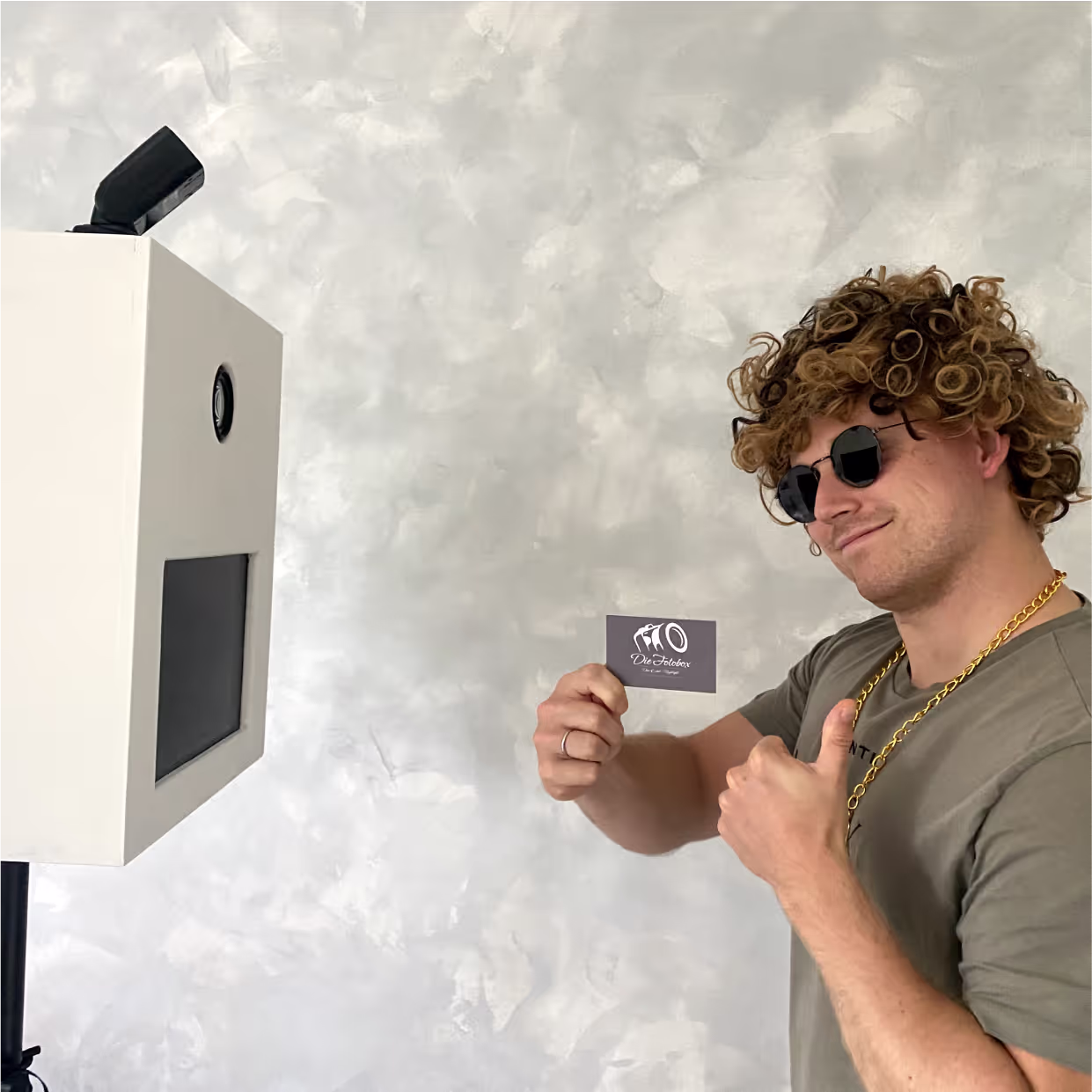 Eine Person mit eine Perücke auf dem Kopf steht neben einer Fotobox und zeigt mit einer Hand die "Daumen hoch" Geste und mit der anderen Hand eine Visitenkarte.